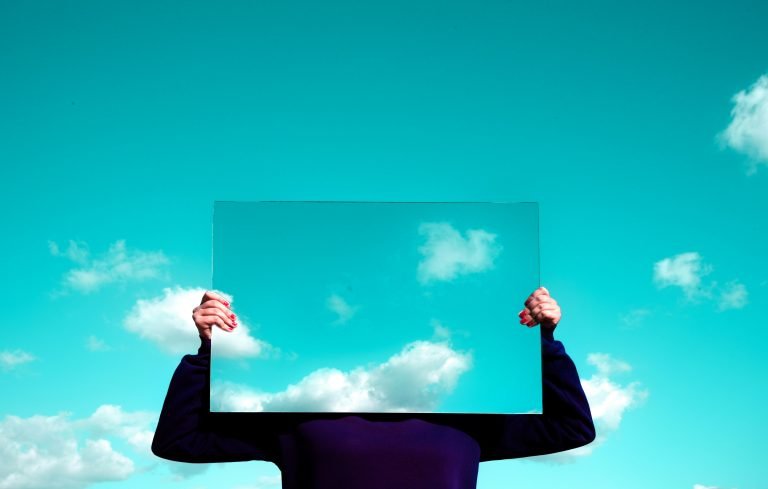Is your marketing still working for you? Let's pause and reflect - image of a person in a purple jumper holding up a mirror reflecting aqua blue sky with a few fluffy white clouds